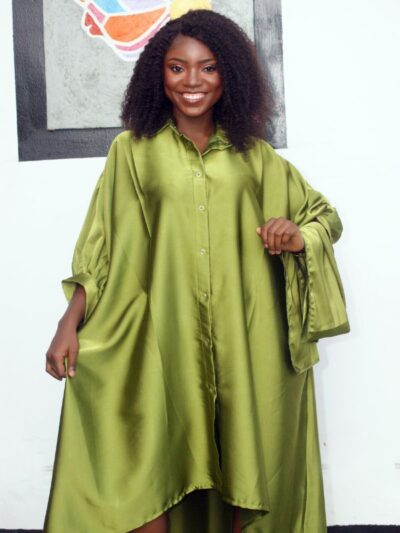 Woman wearing olive green hi-low satin shirt dress with wide sleeves, smiling in front of an African-inspired wall art